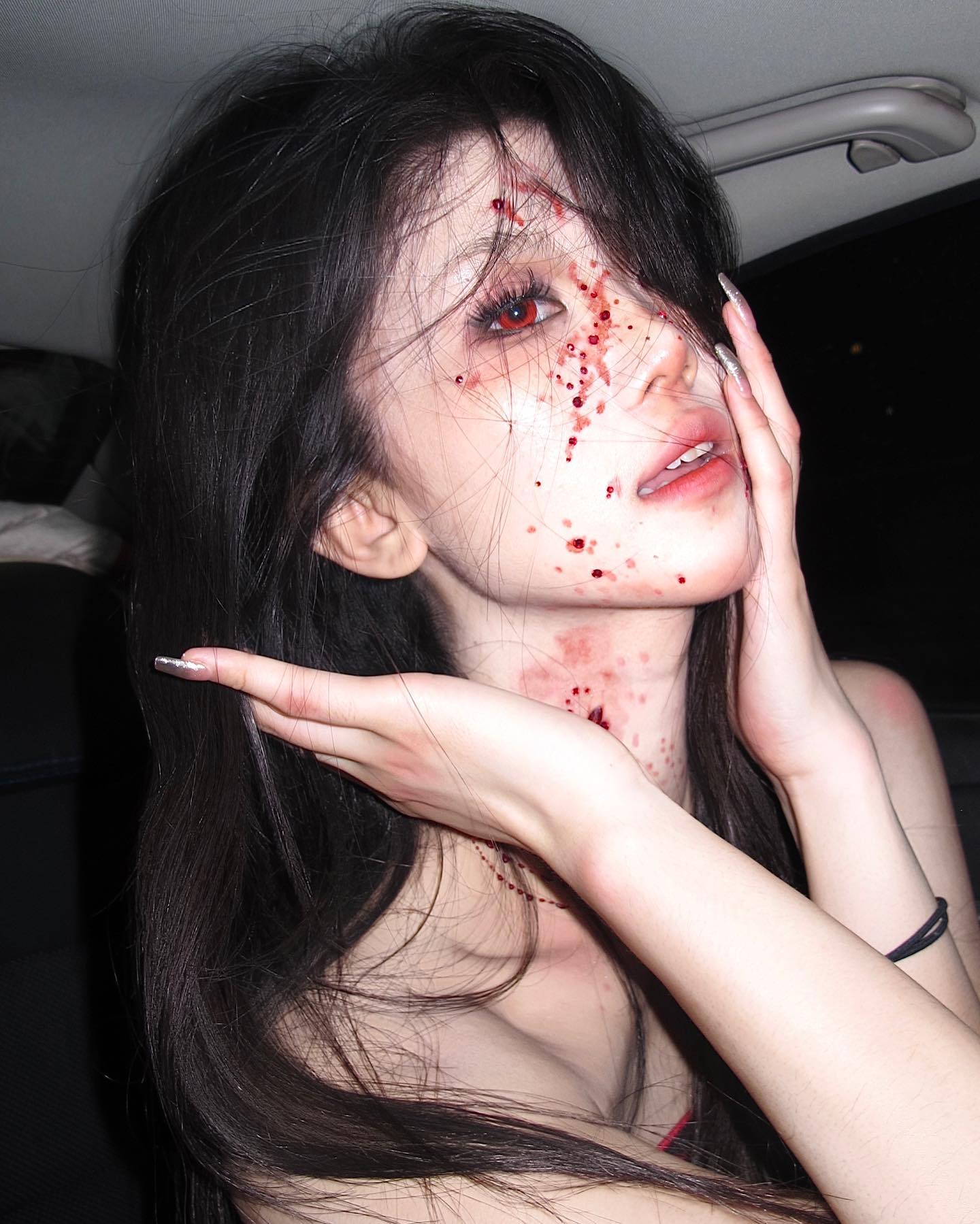 a young woman poses for a picture, showcasing her intricate halloween makeup. she is dressed as a vampire, with fake blood splattered across her face, neck, and chest. her makeup includes pale skin, dark eyes with red contacts, and bloody fangs. her long black hair cascades down, further adding to the eerie effect. the setting appears to be the backseat of a car, with dim lighting adding to the overall spooky ambiance.