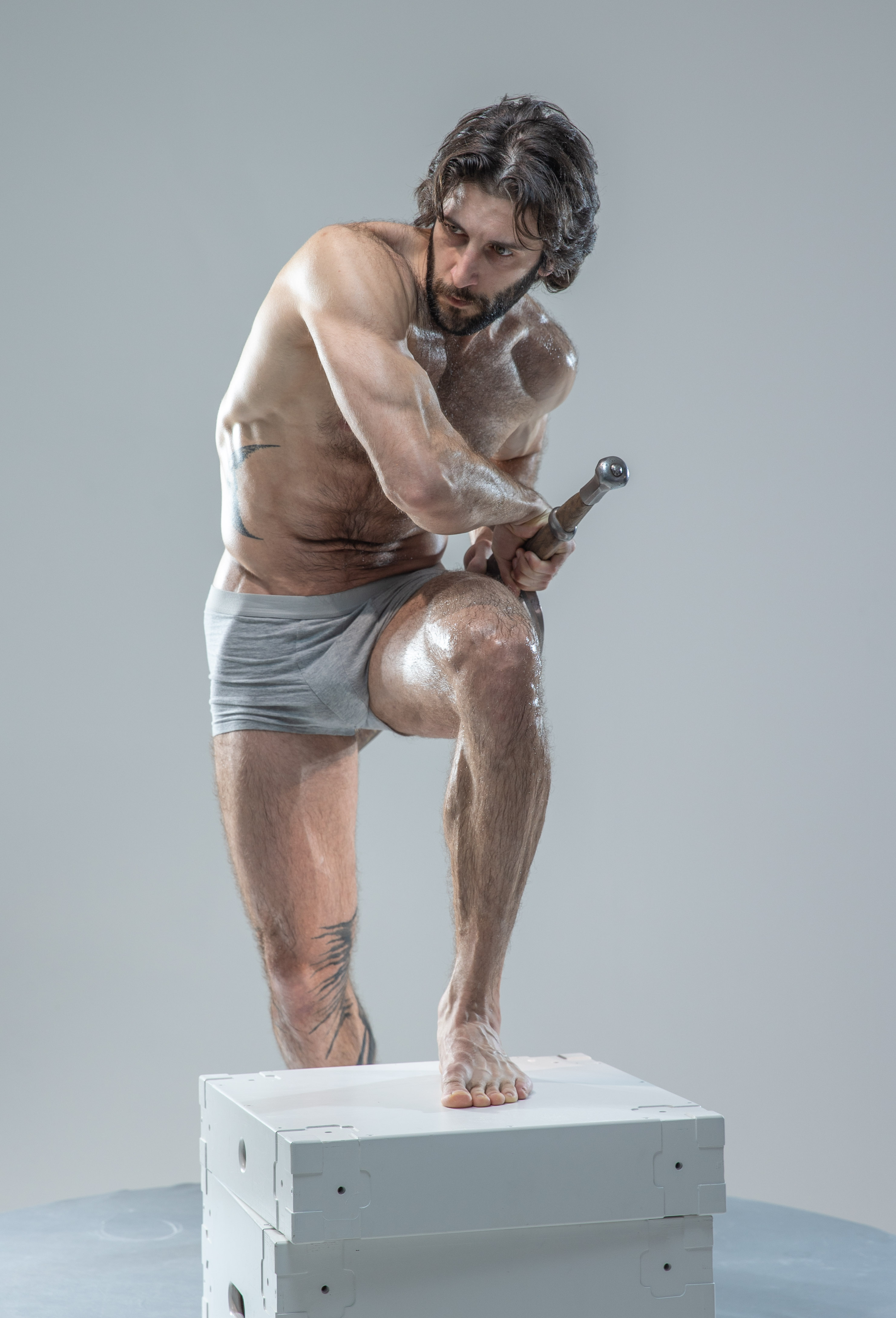 a muscular, shirtless man with dark hair and beard is standing on a white cube in a studio. he is holding a hammer and is covered in sweat. the background is a light gray.