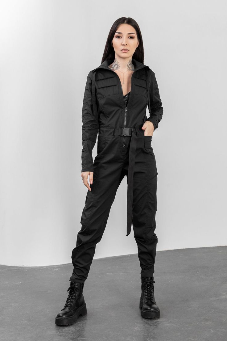 a fullbody studio shot of a woman modeling a black jumpsuit. the jumpsuit features a hooded neckline, long sleeves, cargo pockets, a zipper closure, and a belt. the model is wearing black combat boots and has a neutral expression. the background is plain white.  the overall style is dark, edgy, and contemporary.
