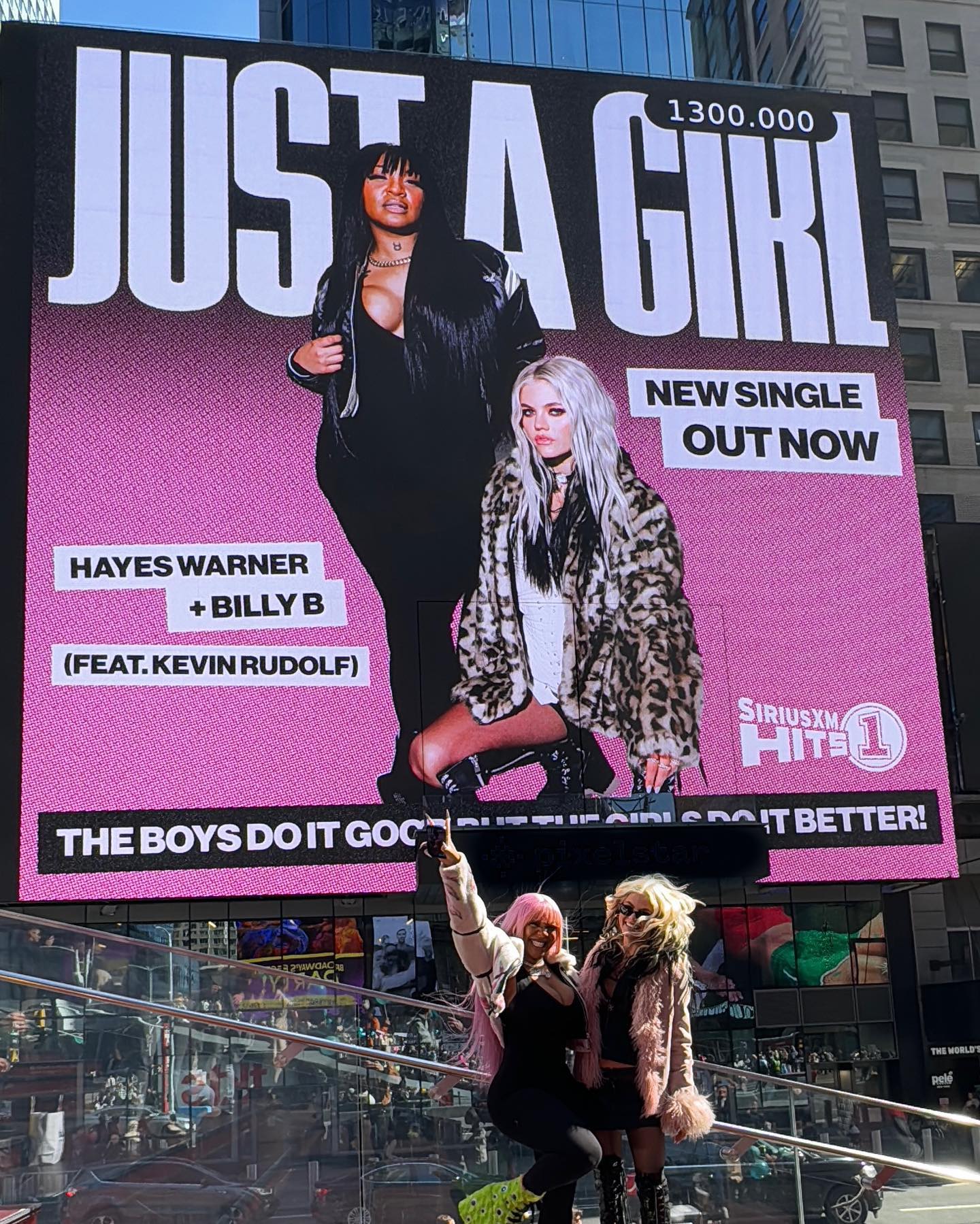 the image shows a billboard in times square, new york city advertising the new single "just a girl" by hayes warner, billy b, and featuring kevin rudolf. the billboard features two women, presumably hayes warner and billy b. below the billboard, two women, one with pink hair and green shoes and the other with blonde hair, are posing for a picture. the billboard also advertises siriusxm hits 1.