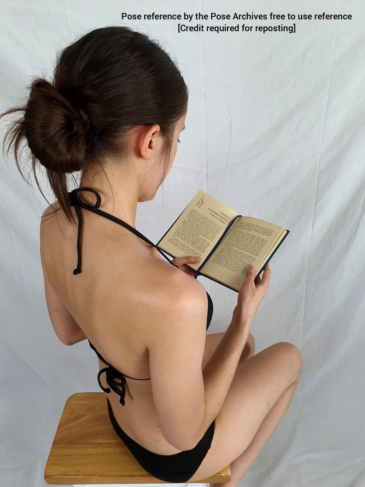 a woman with dark hair in a bun, wearing a black bikini top and bottom, sits on a wooden stool reading a book.  the perspective is from behind and above.  the background is a white wall.```