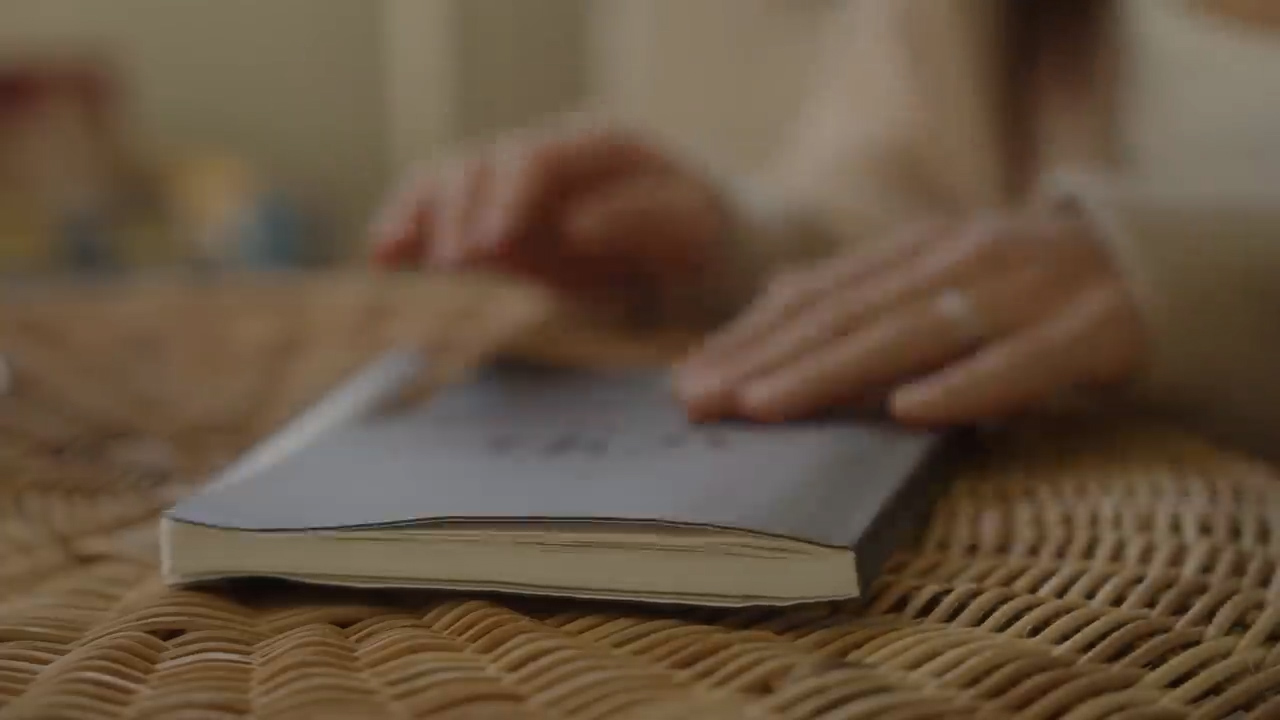a closeup shot of a woman's hands gently resting on a book placed on a wicker table. the soft lighting and neutral color palette create a sense of tranquility and peace.