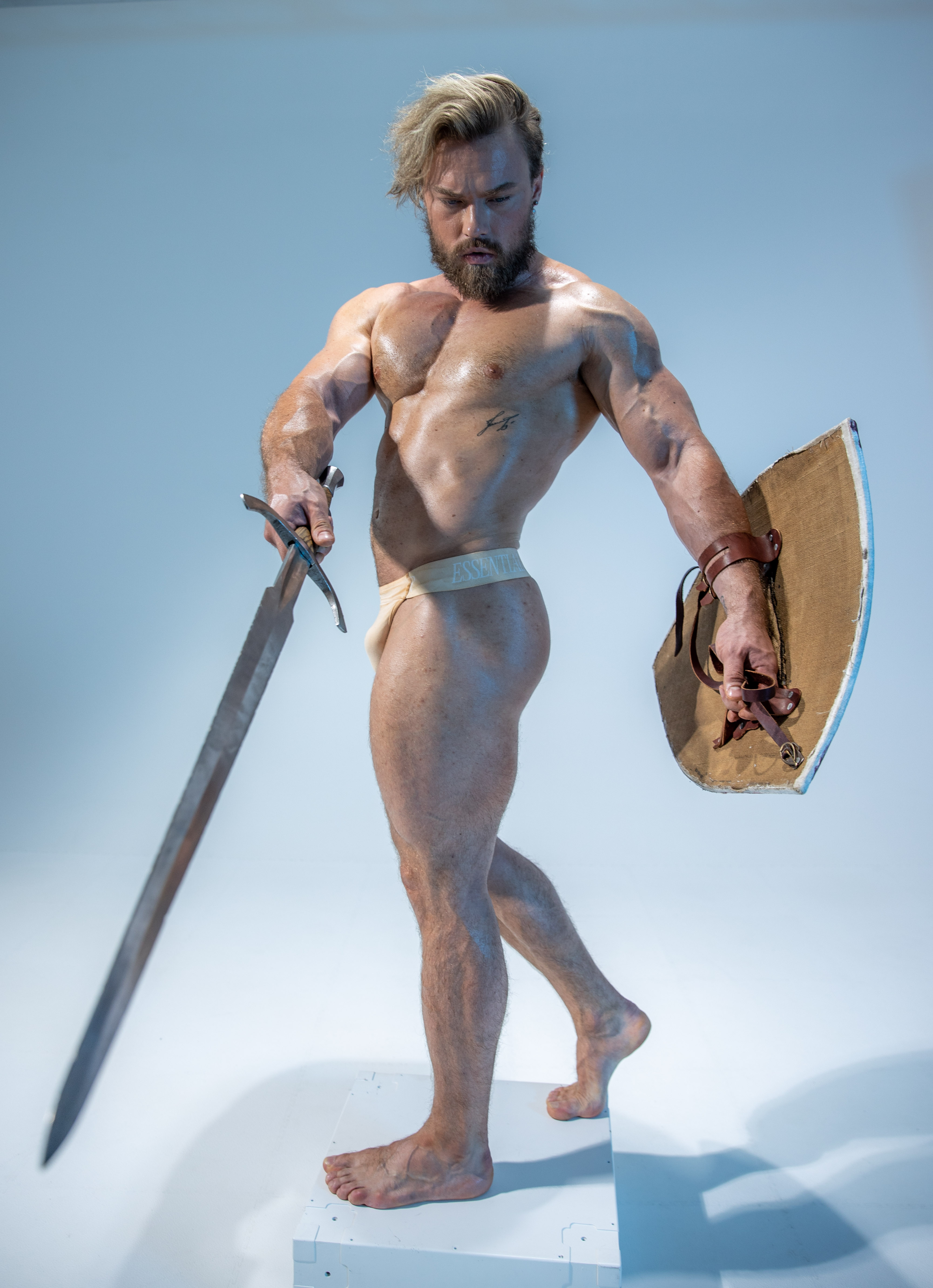 muscular, barechested man stands in a warrior pose against a light blue studio backdrop. he holds a sword in his right hand and a round shield in his left, both poised for action. he is lit artificially, likely with studio lighting, and the camera angle is low, emphasizing his powerful physique. he stands on a white platform. his attire is minimal, consisting only of a simple loincloth, further emphasizing his strength and warriorlike appearance.