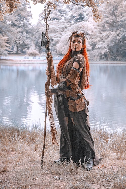 full body shot of a woman dressed in a fantasy costume, possibly a druid or witch, standing by a lake in a forest. she has long, red hair, and is wearing a brown and black outfit with various accessories, including a staff. the setting appears to be autumn or winter, with muted colors and a slightly cold atmosphere. the woman is looking towards the viewer with a serious expression. the overall style of the image suggests a  fantasy or pagan theme.
