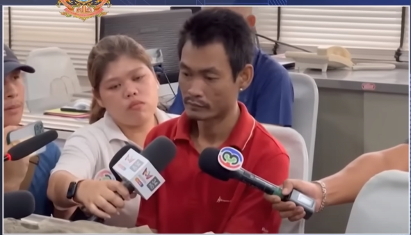 a man in a red shirt is being interviewed by a group of reporters. he is sitting at a desk and there is a woman in a white shirt standing next to him. there are several microphones in front of him. the image appears to be a still from a news broadcast or press conference about a crime.