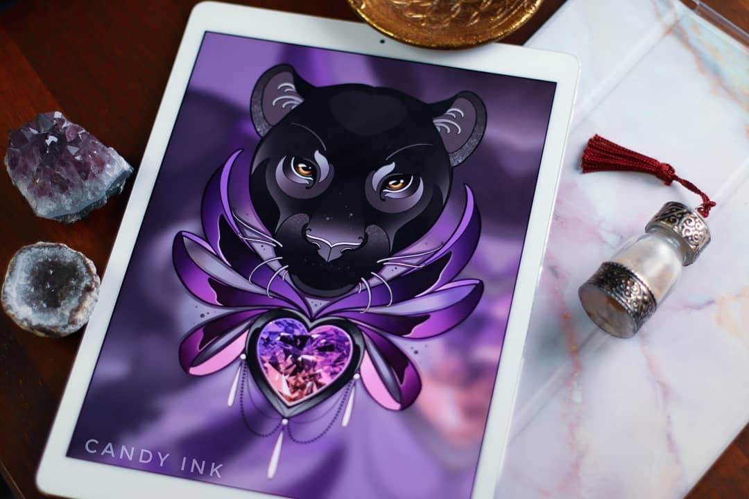 the image showcases a digital art piece of a black panther head design with purple wings stemming from behind. the panther is centered above a jewelencrusted heart. the background is a gradient purple and the art is displayed on a tablet surrounded by crystals, a tray, and a small jar.  the artist's watermark, "candy ink," is visible at the bottom of the image.