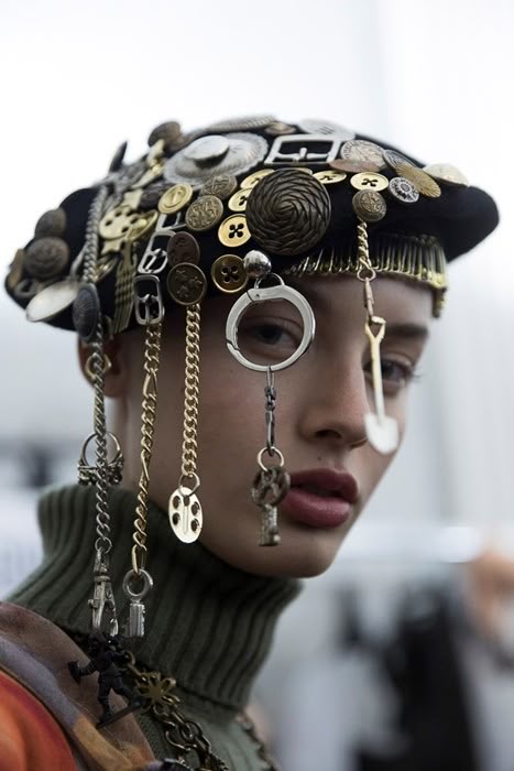 a portrait shot of a woman wearing a unique and extravagant hat adorned with various buttons, keys, chains, and other trinkets. the hat is densely covered with a mix of gold and silver colored metal ornaments, including vintage buttons, small keys, buckles, and coins. chains dangle from the hat, accentuating the eccentric style. the woman has a serious expression and wears dark lipstick, enhancing the high fashion and artistic nature of the image. she is also wearing a green turtleneck sweater and necklace with similar metallic ornaments. the background is blurred, drawing focus to the woman and her striking headwear.
