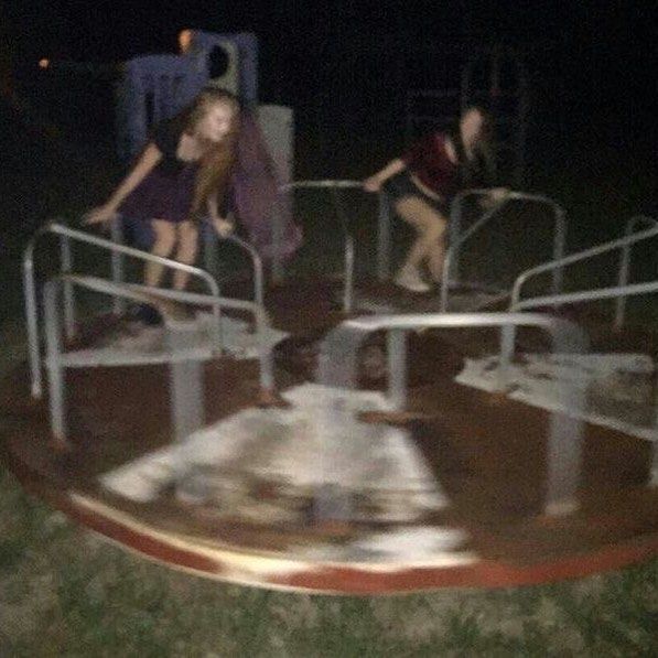 two girls are on a spinning merrygoround in a playground at night. the motion of the ride is blurred. there is other playground equipment in the background.