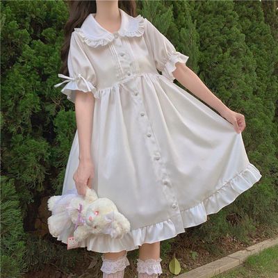 a woman in a white lolita fashion dress standing in a garden. the dress has a peter pan collar, ruffled hem, and button front.
