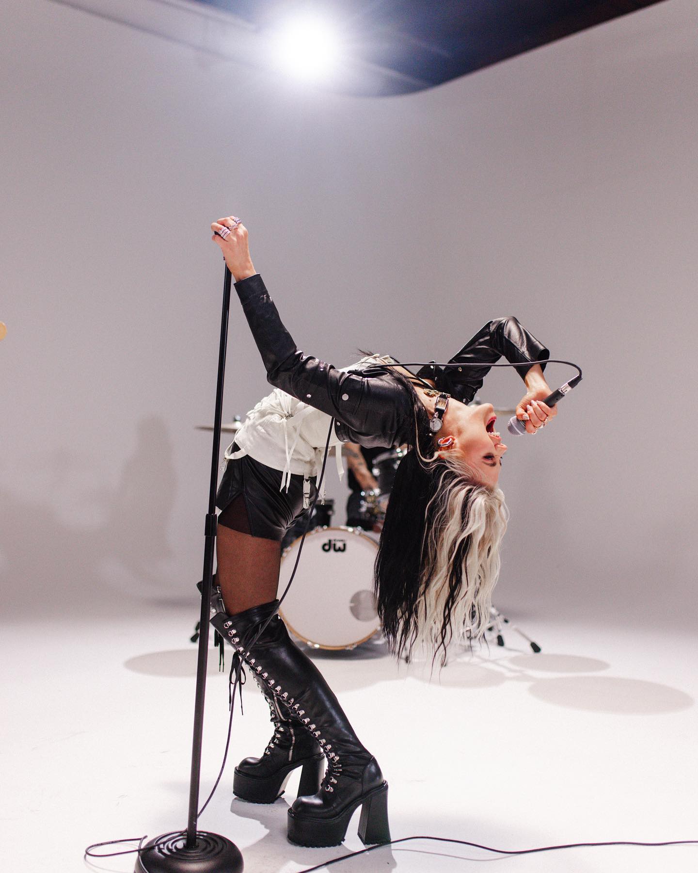 a woman in a black leather jacket, shorts, and platform boots is bent backwards while singing into a microphone. she has black and white hair and is on a stage with drums in the background. she is performing music.