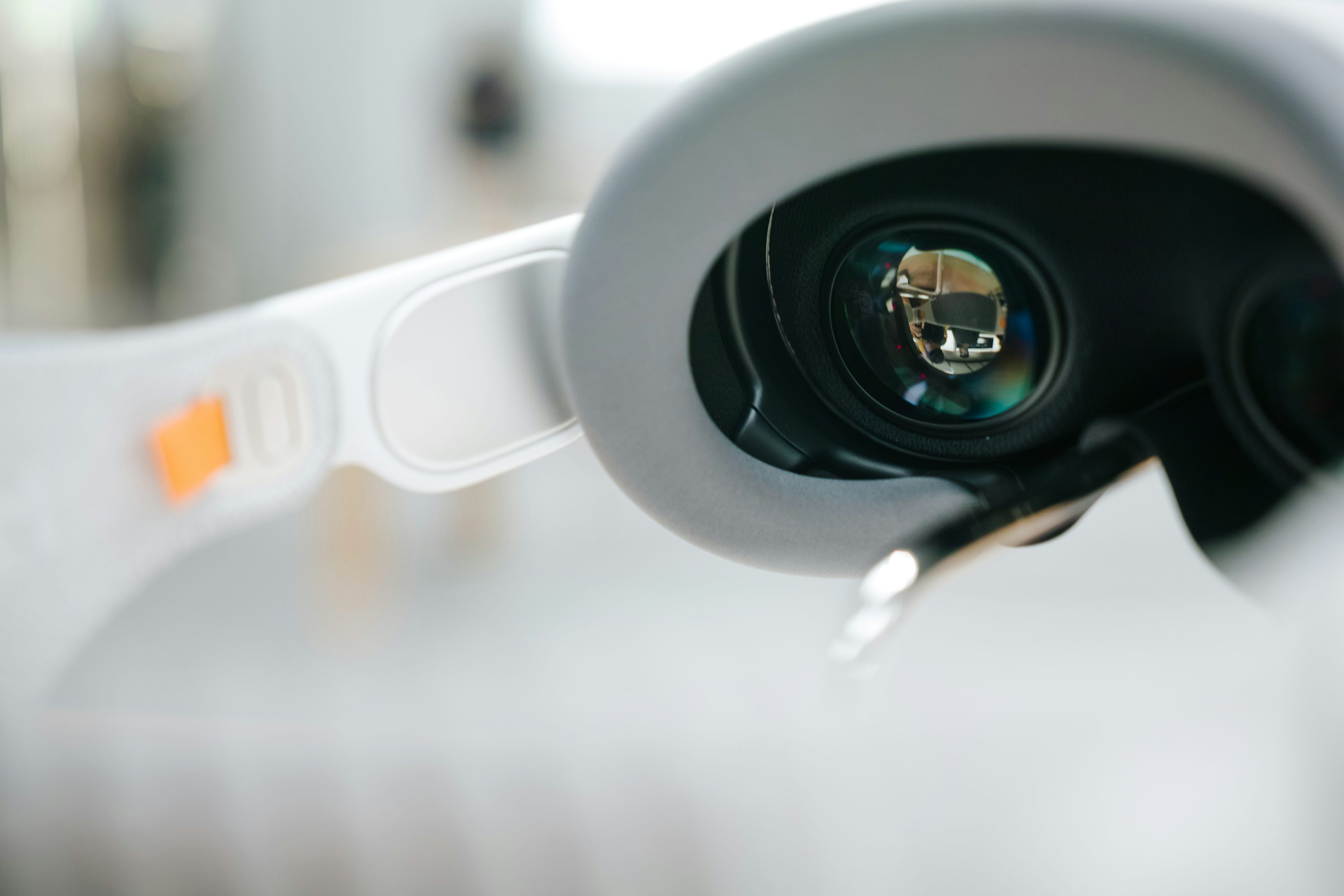 a closeup shot of a white and gray vr headset with its lenses prominently displayed, reflecting the surrounding environment. the headset appears modern and futuristic, with an orange detail on the side. the focus is on the lenses, highlighting the immersive technology of virtual reality.