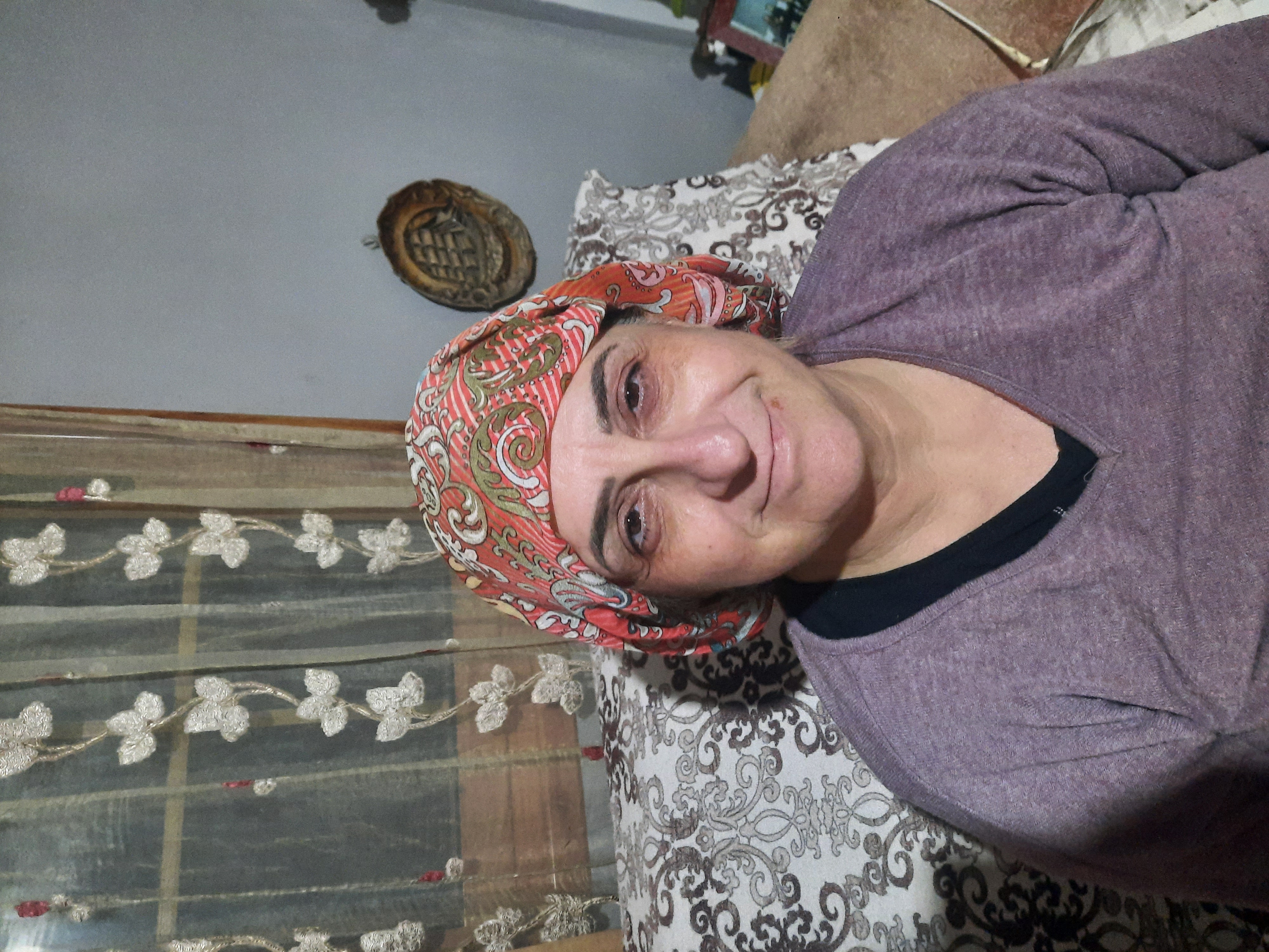 a portrait of a woman wearing a patterned headscarf and a purple vneck sweater. she is smiling and sitting on a couch indoors. behind her, there is a curtain with a floral pattern and a wall hanging on the gray wall.