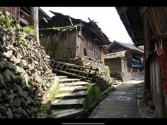 this image captures a narrow pathway winding through a historic chinese village. the path is paved with stone, and steps lead upwards to the left. on either side of the path are traditional houses, constructed from wood and stone. the buildings appear weathered and aged, hinting at a rich history. a stone wall and a makeshift wooden fence line the path. sunlight bathes the scene, casting shadows and highlighting the textures of the stone and wood.
