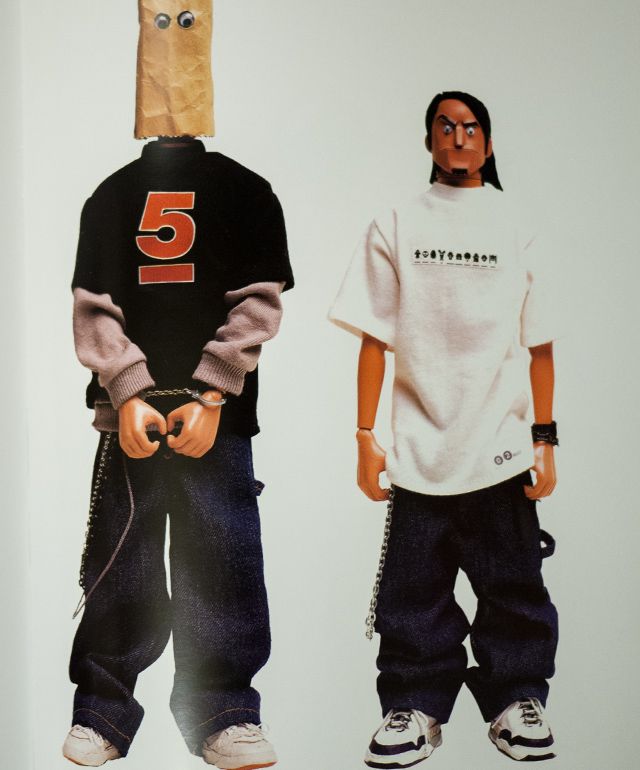 two male dolls are dressed in casual street wear. the doll on the left is wearing a brown paper bag on his head, a black tshirt with a red number "5", gray hoodie, blue jeans, chains, white sneakers and has handcuffs on. the doll on the right is wearing white tshirt, a  gray hoodie, blue jeans, a black belt with chains, white sneakers, a black headband and sunglasses. they both have white skin, brown hair and brown eyes.  the dolls are standing on a white background.