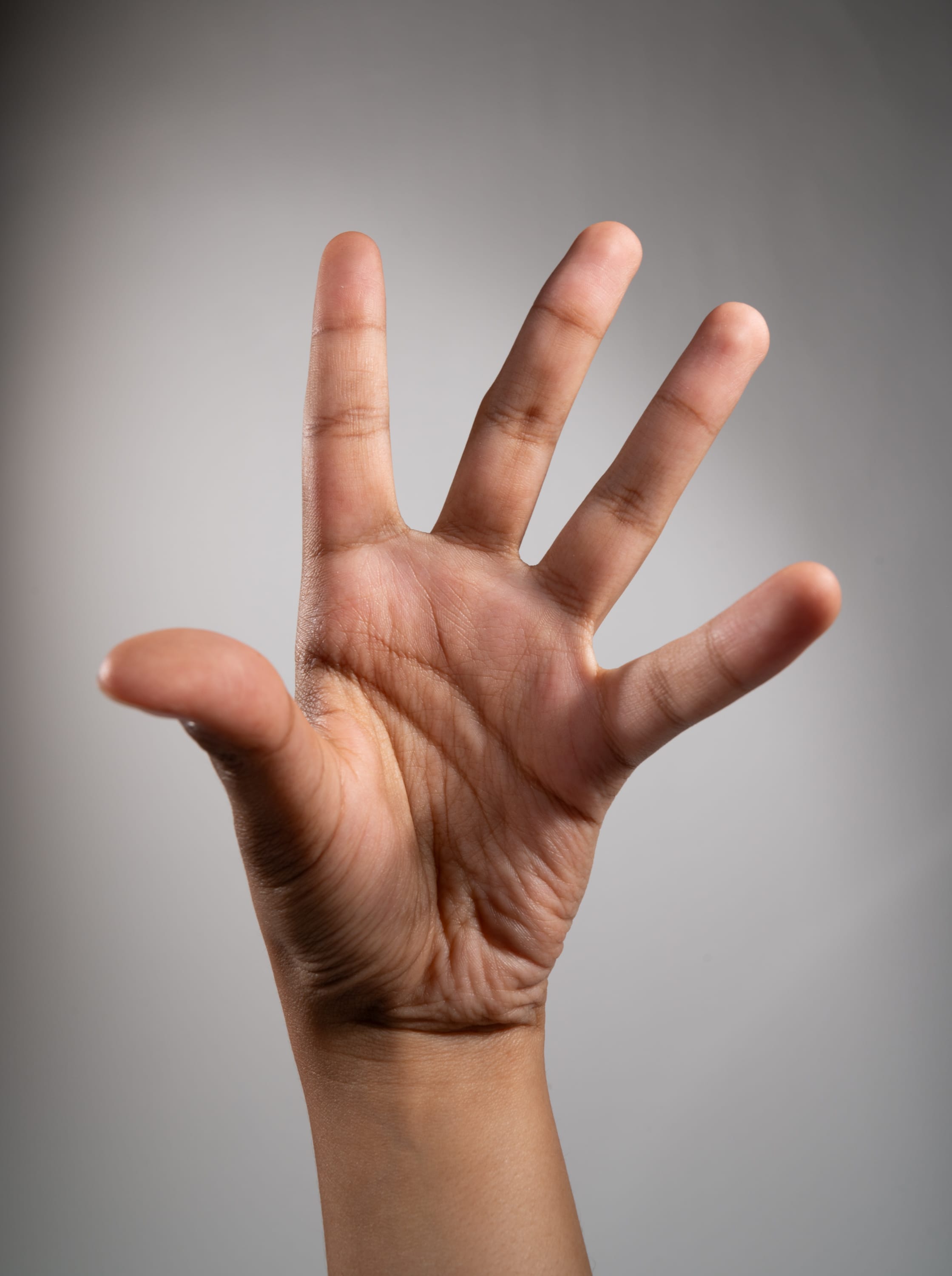the image shows a hand with an open palm and fingers extended, set against a neutral gray background. the hand is positioned vertically, with the palm facing outward. the lighting is soft and even, highlighting the details of the skin and hand structure. the image focuses on the hand's shape, gesture, and the subtle variations in skin tone.