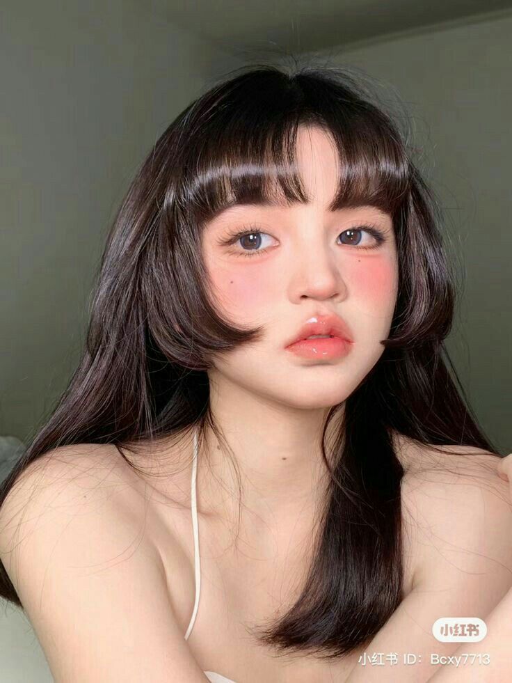 closeup portrait of a young asian woman with dark, straight hair styled in a bob with blunt bangs. she has rosy cheeks, soft makeup, and is looking directly at the camera.  the background is blurred and out of focus, drawing attention to the subject. the overall aesthetic is soft, delicate, and charming.  a small watermark is visible in the lower right corner.