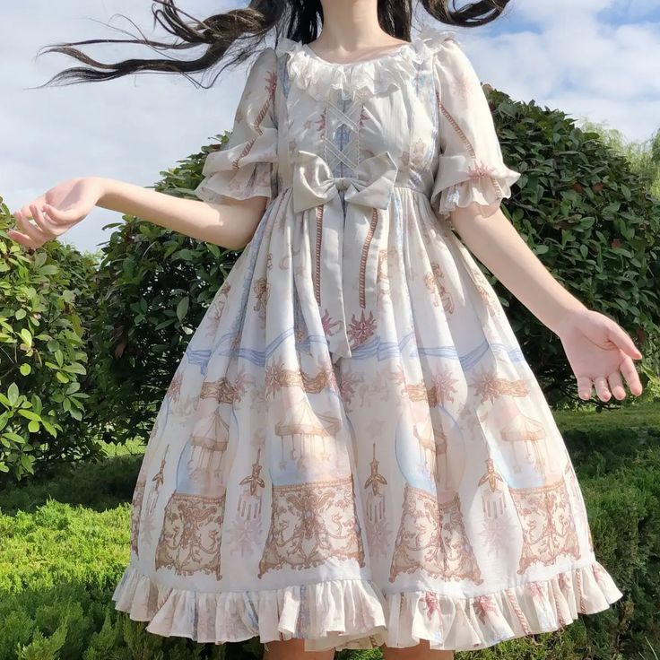 a young woman wearing a flowing white lolita dress with a blue and brown pattern stands in a garden setting.  the dress features puffed sleeves, a lacetrimmed square neckline, and a ruffled hem. she is posing with her arms outstretched, and her face is not visible. the lighting is bright and sunny, highlighting the details of the dress and the greenery of the garden.