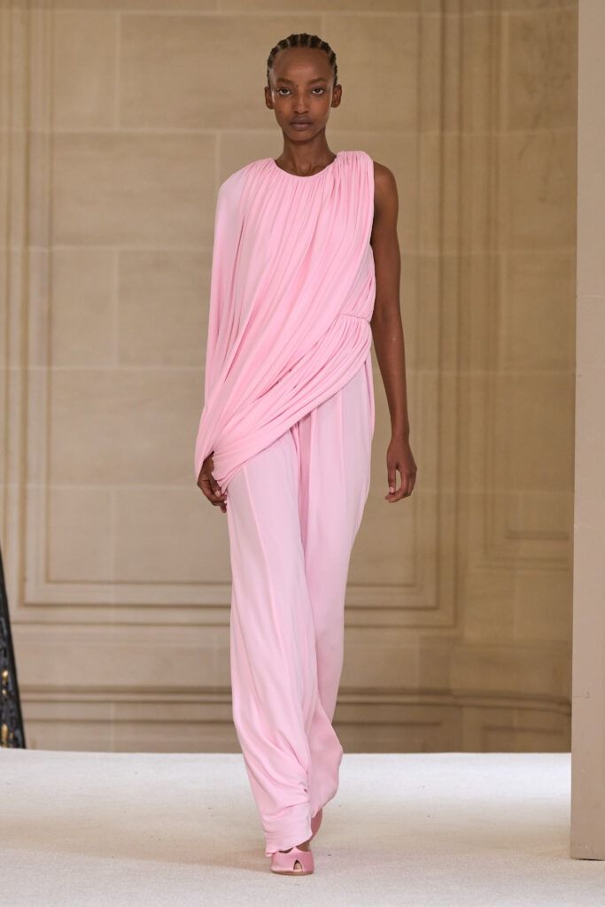a black woman with cornrow braids is walking down a runway wearing a pink, oneshoulder jumpsuit. the top of the jumpsuit is draped and the pants are widelegged. she is wearing pink shoes. the background is a beige wall.