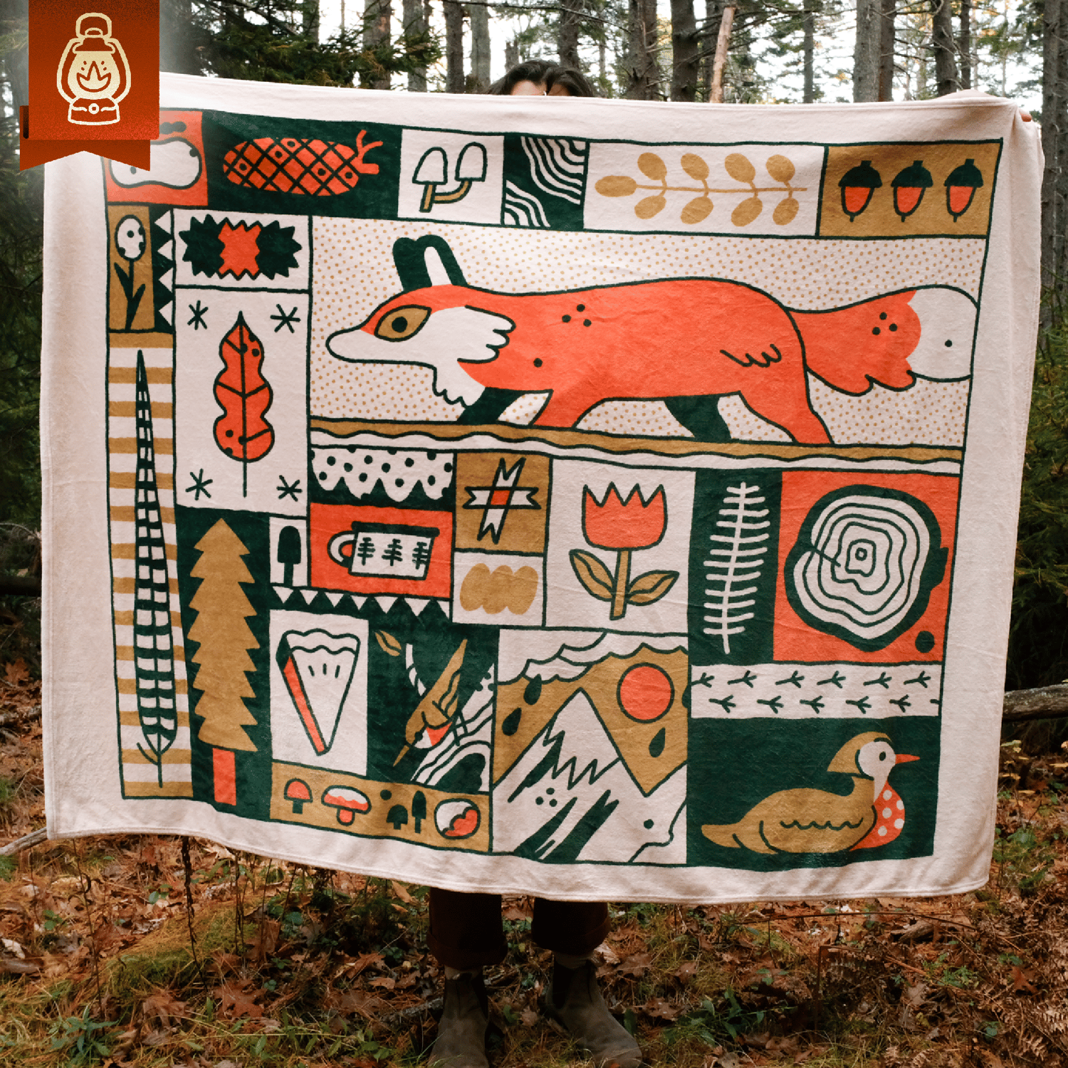 a person is holding up a large, creamcolored throw blanket featuring a colorful and whimsical illustration of woodland creatures and nature scenes. the design is primarily composed of geometric shapes and bold colors, including oranges, greens, and golds.  a prominent feature is a stylized orange fox walking across the central portion of the blanket. other elements include mushrooms, acorns, a bird (possibly a wood duck), leaves, and a stylized mountain range.  the blanket's fabric appears soft and textured. the background is a forest setting, suggesting the blanket's intended use for outdoor activities. the overall style is reminiscent of vintage or folk art designs.