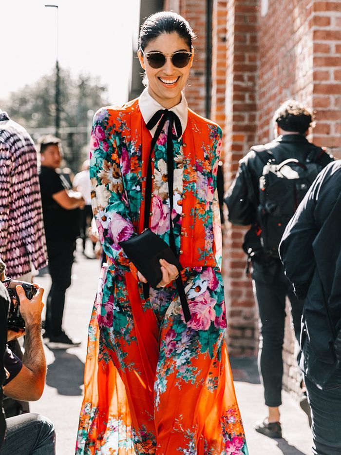 a young woman in a trendy red and orange floral dress and black sunglasses walks confidently down a city street. she is smiling and looking straight at the camera.  she is elegantly dressed and her style is both chic and casual. the background of the photo is a brick wall and blurry people.