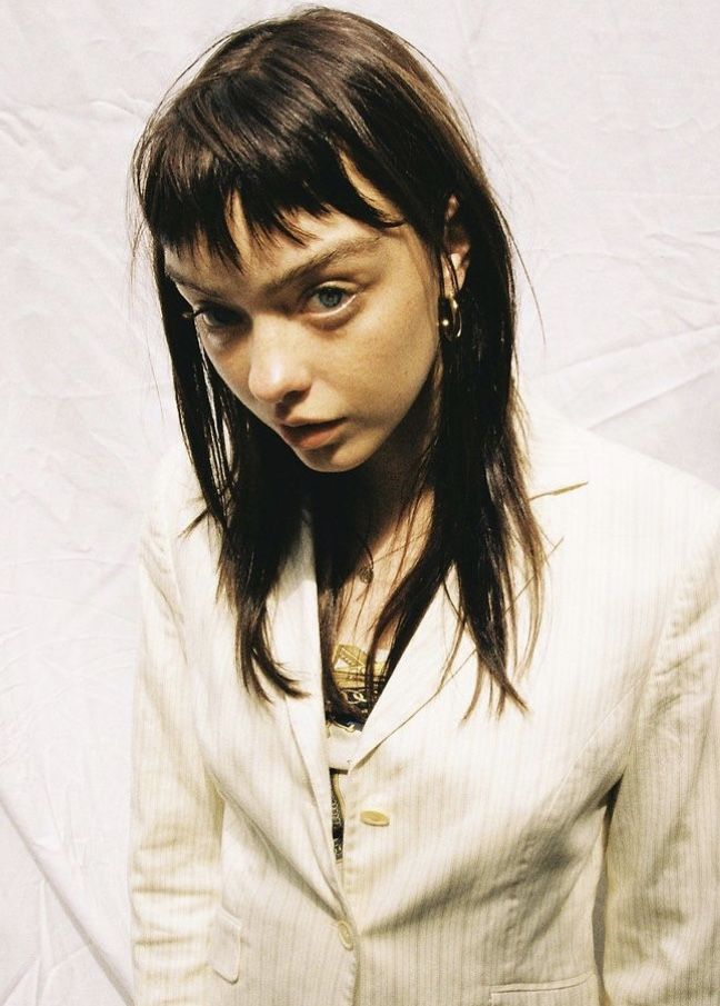 closeup portrait of a young woman with straight dark brown hair and bangs, wearing a light beige pinstripe blazer. she is looking directly at the camera with a serious expression. the background is a light, textured fabric. she has gold hoop earrings. the image is welllit and appears to be a professional studio shot.