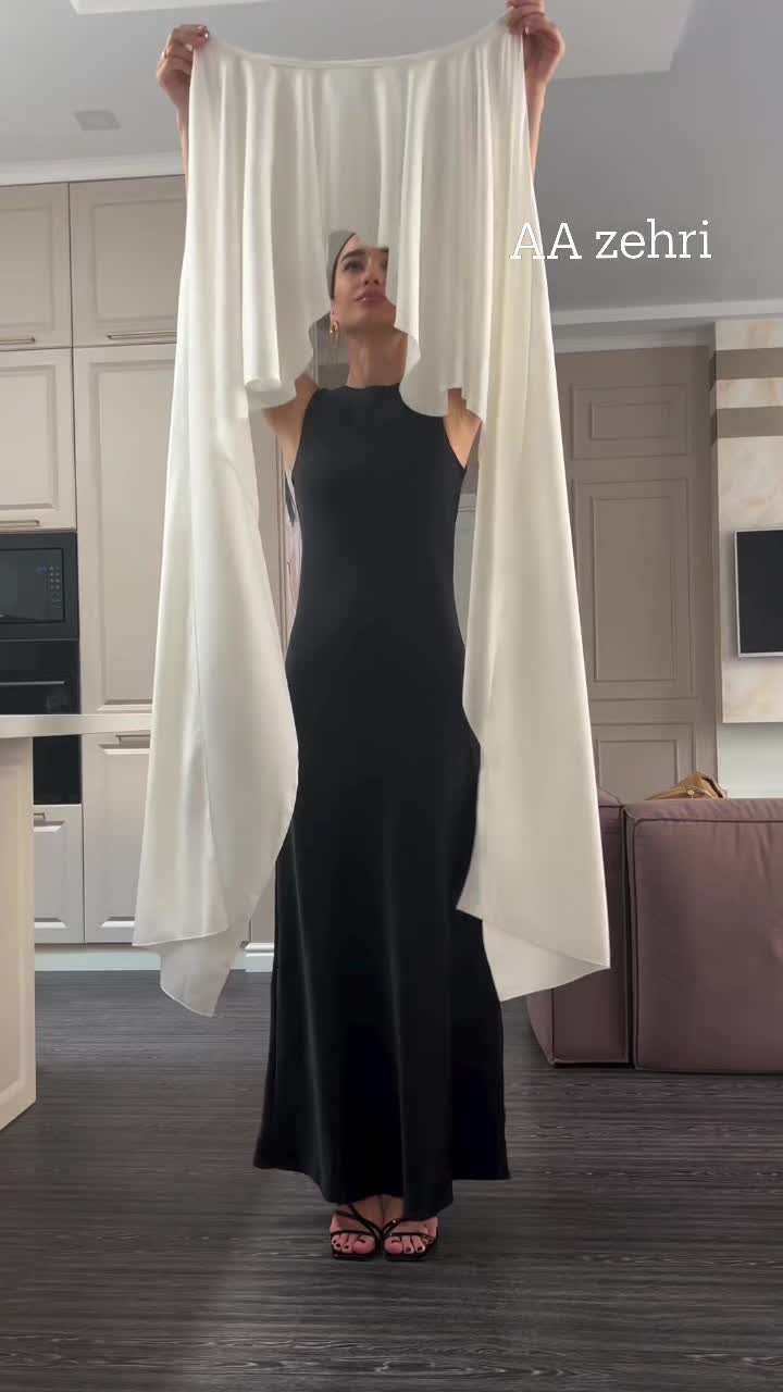 a woman is standing indoors, wearing a black dress. she is holding a large piece of white fabric up over her head, with the fabric draping down on either side of her. the background includes a kitchen area with cabinets and a counter.