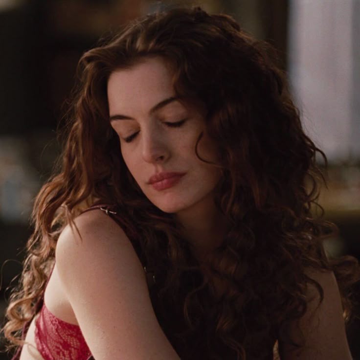 a portrait of actress anne hathaway with long, curly brown hair, wearing a red lace dress. she is looking down with her eyes closed, appearing pensive or serious.