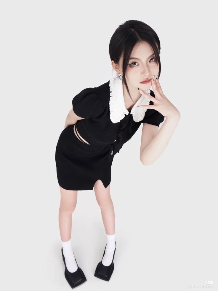 a young woman with black hair is wearing a black dress with a white collar. she is posing in a studio with a white background. she is looking at the camera and has a serious expression on her face. she is wearing white socks and black shoes.