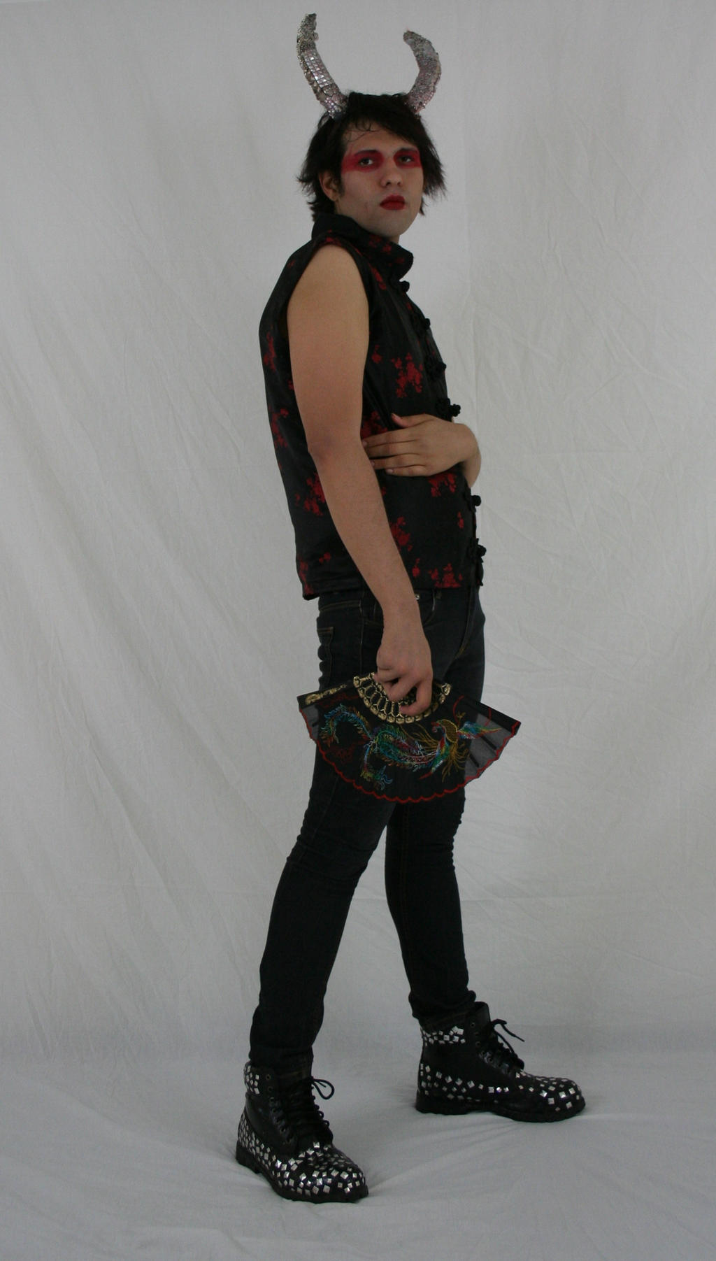 full body shot of a person in a black and red chinesestyle top, black jeans, and studded boots.  they are wearing silver devil horns and red makeup. they are holding a hand fan. the background is white. the image is a studio shot.  the person appears to be a model posing for a fashion or cosplay photograph.