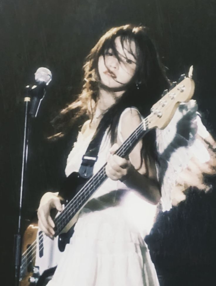 the image shows a young woman with long dark hair playing an electric bass guitar on a stage.  she's wearing a white dress and appears to be performing a concert. the background is dark and somewhat blurry, giving the photo a grungy, vintage feel. the lighting is low, focusing primarily on the woman and her instrument.  the overall aesthetic is dark, moody, and evokes a feeling of indie or alternative rock. the woman's expression is intense and focused.