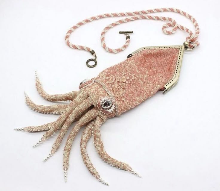 the image shows a beaded squid purse on a white background. the purse is mostly pink and white, resembling a squid with tentacles and pearls at the tips. the eyes are jeweled, and it has a beaded necklace attached with a clasp. the top of the purse has a metal frame, suggesting it opens like a handbag. the overall impression is a unique and artistic fashion accessory.