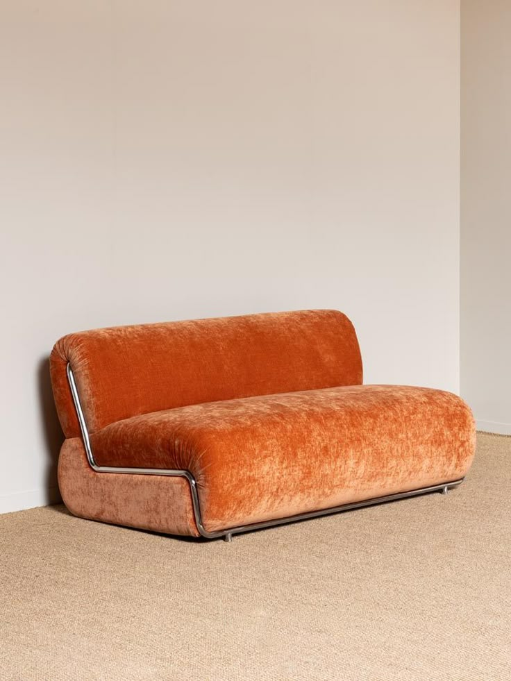 an eyelevel, full studio shot of an orange velvet loveseat with a chrome metal frame. the loveseat is on a beige textured floor, and it's set against an offwhite wall. the loveseat has a rounded back and a rounded seat. the chrome frame curves around the back and seat, and there are small metal legs.