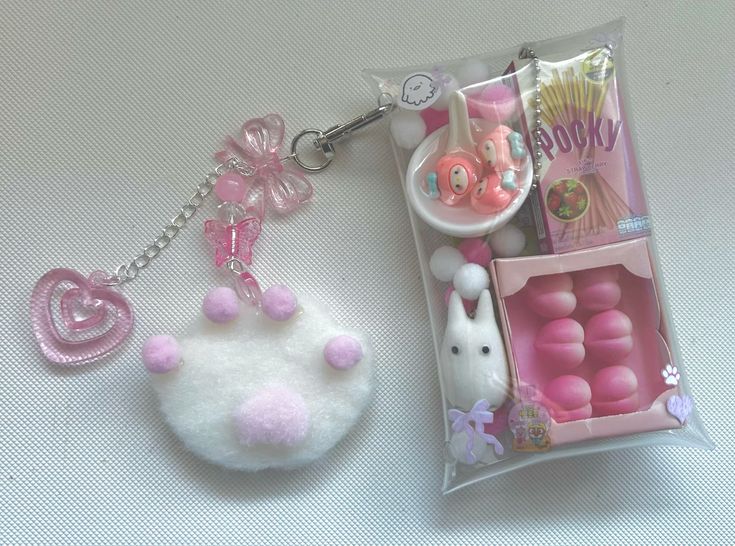 the image shows two keychains. one is a white fuzzy paw print with pink dots and a pink heart and butterfly charm. the other keychain is sealed in a clear plastic package and contains candy, snacks, and a totoro figurine. the snacks appear to be pocky. the color scheme is primarily pink and white.