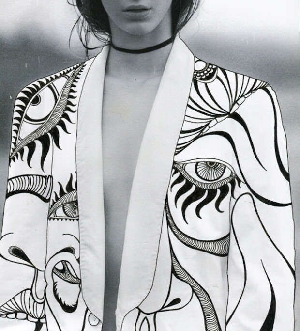 the image is a closeup shot of a woman's torso, showcasing a white blazer adorned with intricate black and white graphic illustrations.  the design features stylized eyes and abstract line work, creating a surreal and artistic effect. the woman is wearing a black choker necklace and her dark hair is partially visible. the overall style is fashionforward and avantgarde. the background is a blurred gray, making the jacket the central focus of the image. the style of the illustration is reminiscent of handdrawn work.