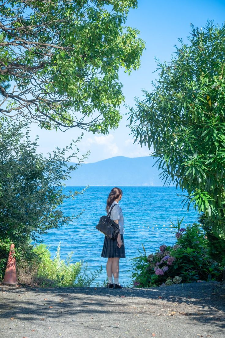 a woman in a skirt stands by a lake, framed by trees. in the distance, a mountain is visible under a bright sky.