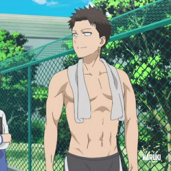 an anime still of a young, muscular man in his swim trunks, with a towel around his neck. he is standing outdoors on what looks like school grounds.  there is a fence and trees behind him. the weather looks sunny. another person's arm can be seen on the left side of the image.