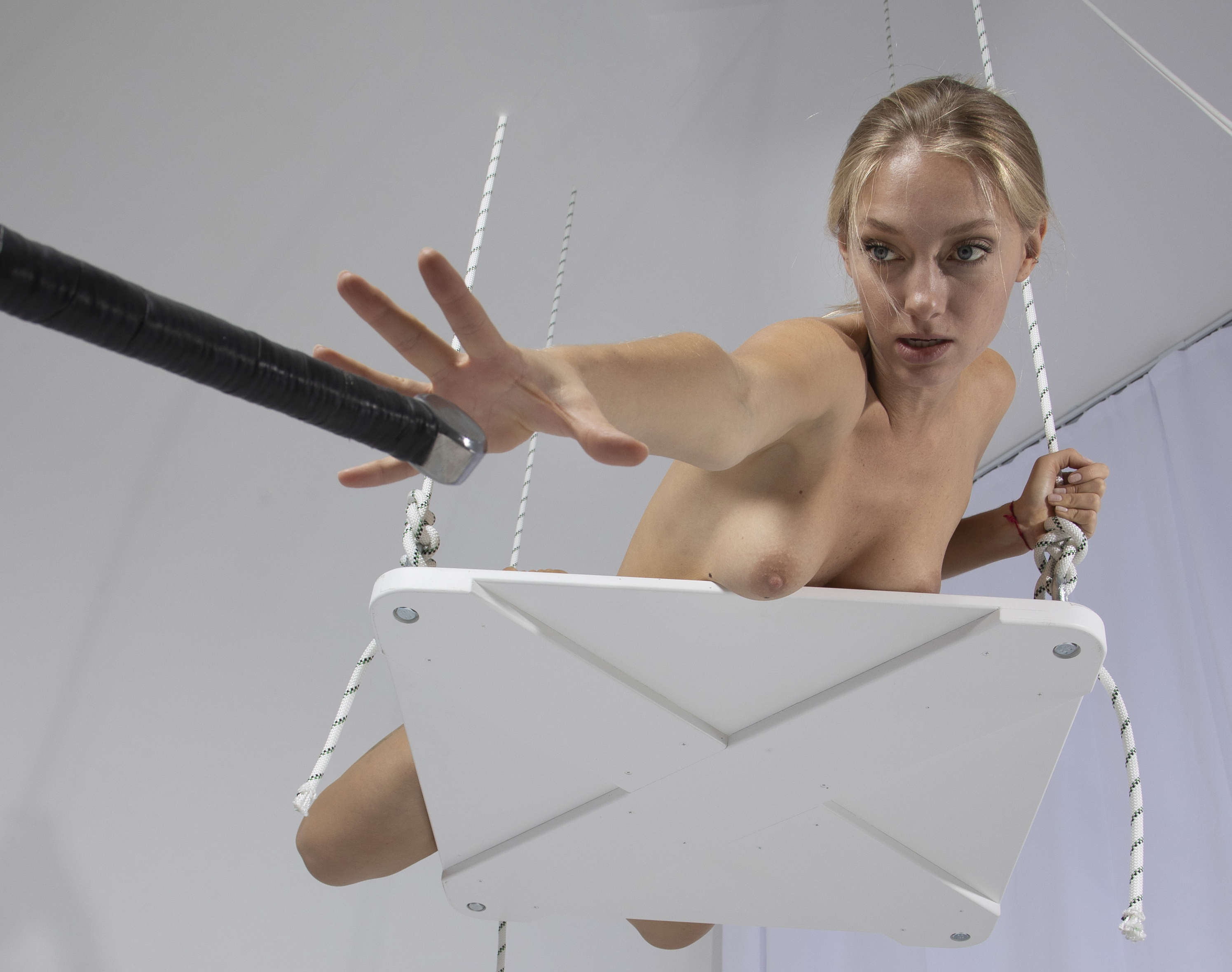 a nude woman is on a white rope swing reaching out to a black pole with a silver tip. the background is white.