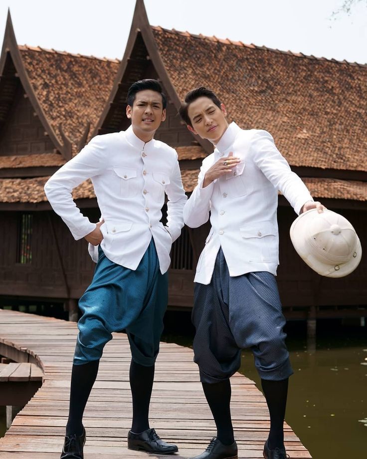two handsome asian men wearing traditional thai clothes are standing on a wooden bridge over a pond in front of a traditional thai building. both men are smiling and look at the camera. they are both wearing white jackets with blue pants. the man on the left is holding a white hat.