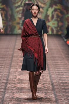 a fashion model walks down the runway wearing a black dress with a red scarf. she is posing for the camera while wearing brown boots. the runway is a dark brown wood floor, with the background blurred and showing green plants.