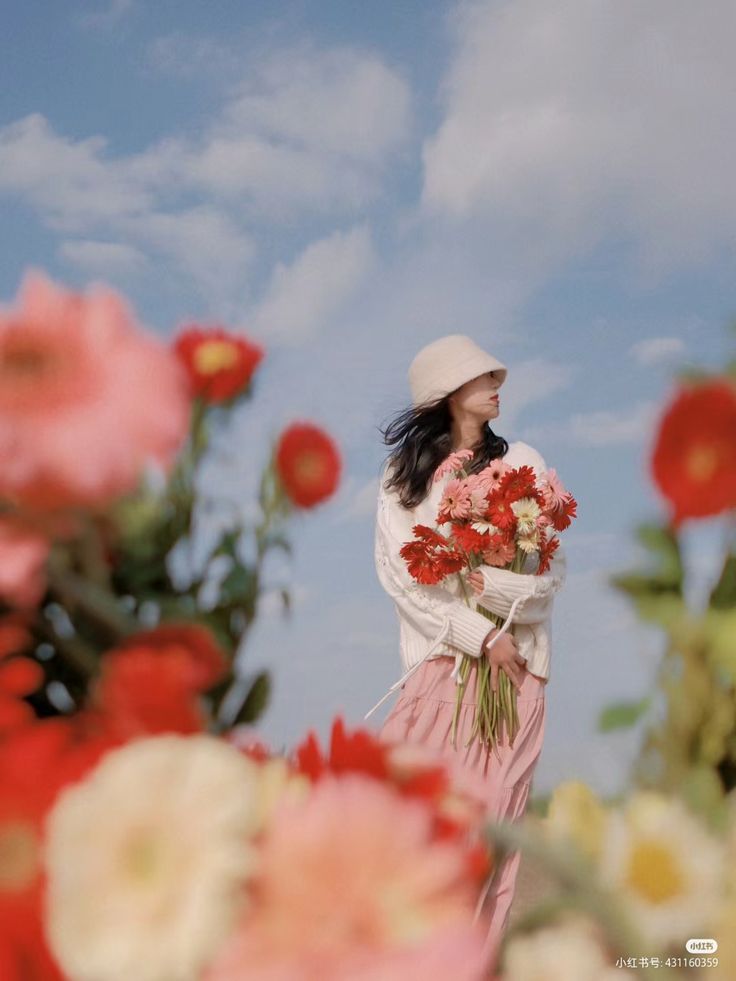 a young woman in a white sweater and pink skirt stands in a field of flowers, holding a large bouquet of gerbera daisies. she is wearing a white bucket hat and looking away from the camera. the sky is blue and cloudy, and the overall mood is serene and joyful.