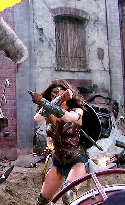 wonder woman in her costume is holding a sword and shield on a movie set. she is wearing a costume and appears to be ready for a fight.  she is standing in front of a large metal structure.  the background appears to be a city scene with buildings and debris.