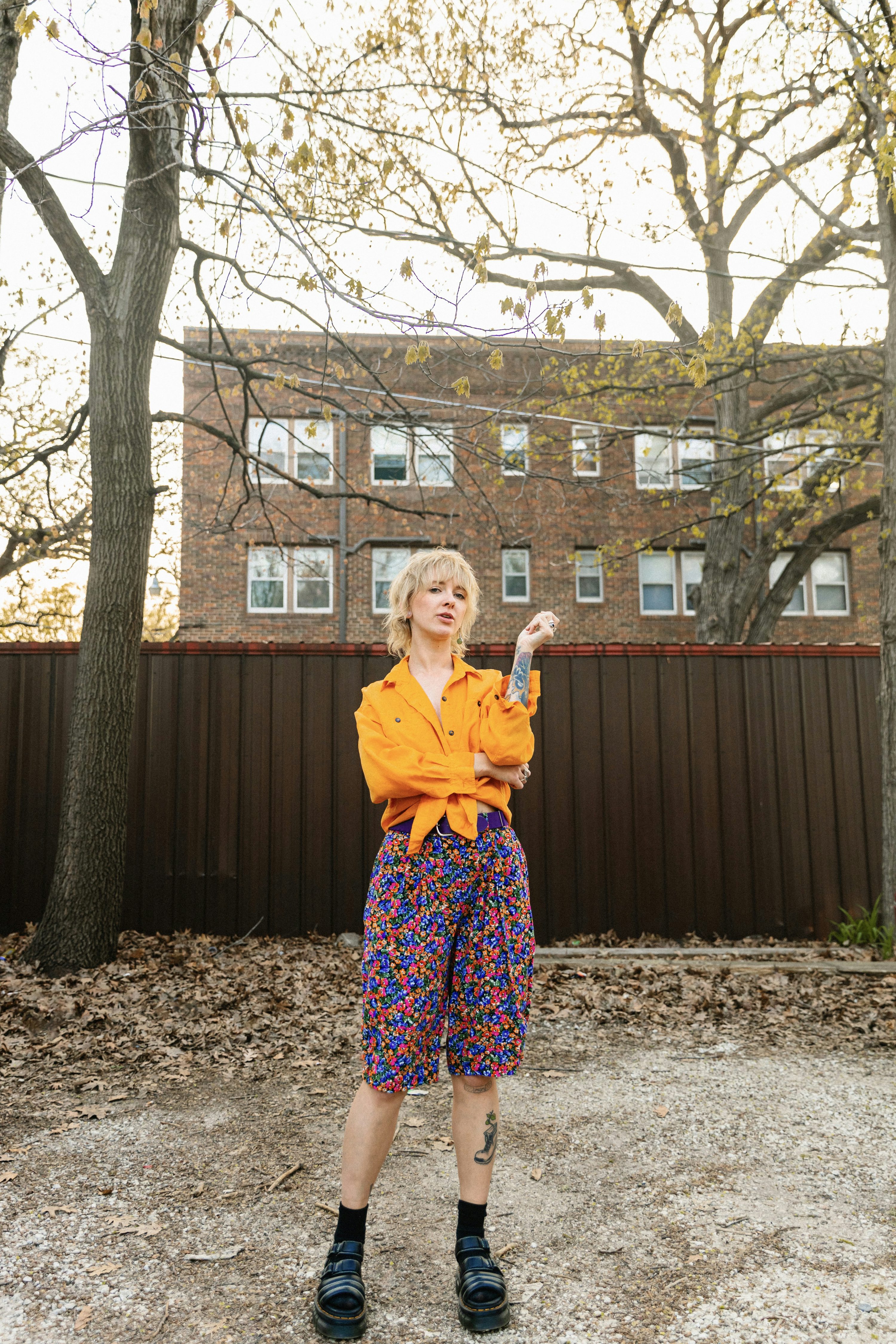 a woman stands in a backyard, wearing a mustardcolored buttonup shirt and colorful floral shorts. she's wearing doc martens and has short blonde hair. a brown fence and a brick building are visible in the background.  the overall style is vintage or retro, possibly 90s inspired.  the setting appears to be springtime, with trees in the background.  the woman has a relaxed, casual pose.