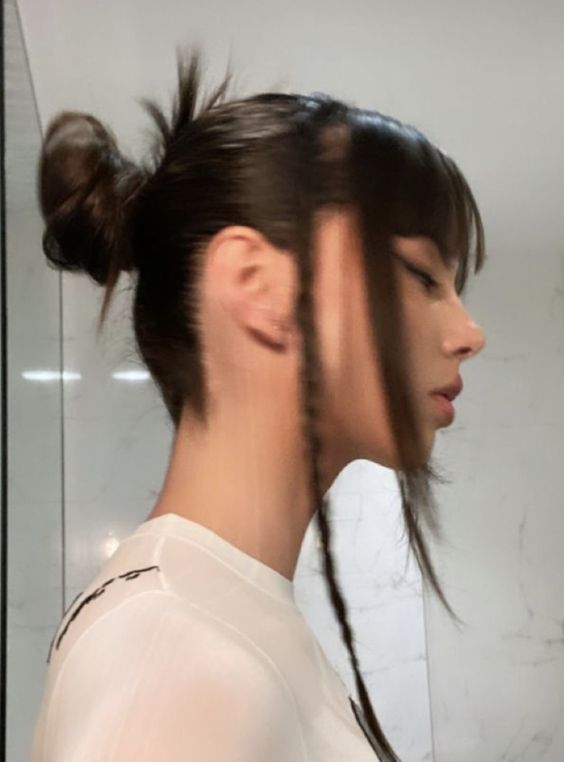 a young woman with a trendy egirl hairstyle featuring a high bun, long faceframing braids, and choppy bangs. she is wearing a white tshirt and is standing in a bathroom with white tiles and a glass shower door. the lighting is natural and the perspective is a side view.