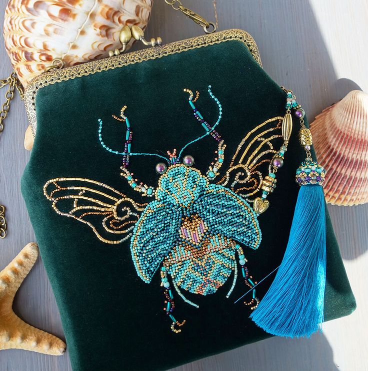 a green velvet purse with a beaded insect design, blue wings, and a blue tassel. the purse has a gold chain and is embellished with jewels. it is sitting on a table with a shell and a starfish.