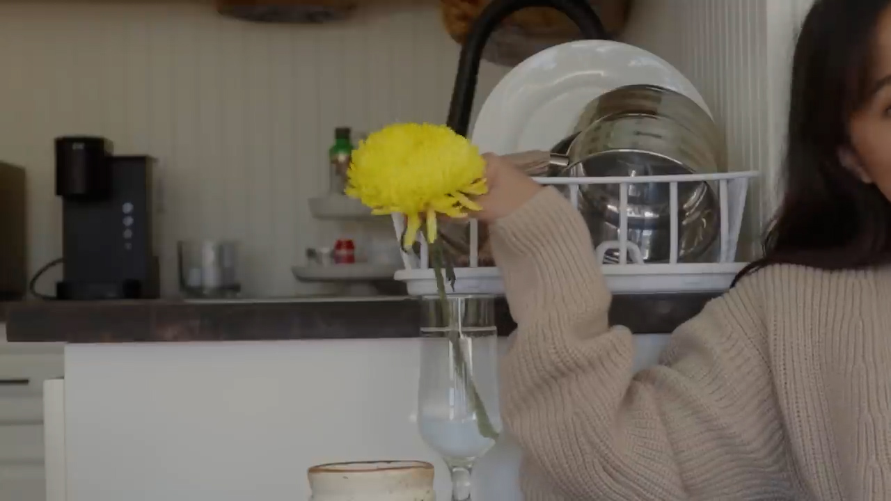 a woman in a beige sweater is standing in a kitchen and reaching up to a white dish rack. she is holding a yellow flower in her hand. there is a coffee maker on the counter behind her. the kitchen is bright and airy, with white cabinets and light wood countertops.