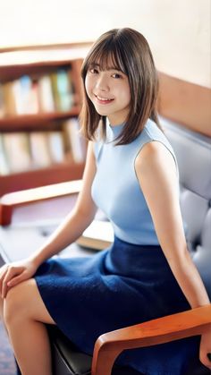 a young woman with shoulderlength dark hair smiles gently at the viewer while sitting on a brown chair. she wears a light blue sleeveless top and a navy blue skirt. her left hand rests on the chair's armrest, while her right hand rests on her thigh. a book lies on the chair beside her. the background features a bookshelf filled with books, suggesting a home library or a cozy reading nook. the image exudes a sense of peace, tranquility, and love for reading. the lighting is soft and natural, creating a warm and inviting atmosphere.