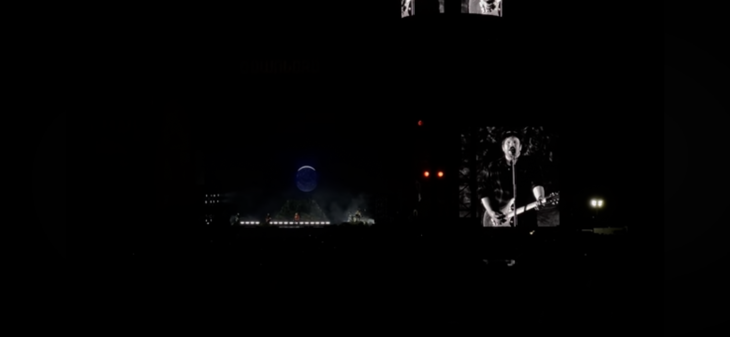 a concert performance at night. the stage is illuminated with bright lights and a large screen is displaying a black and white image of a musician playing a guitar. there are people in the audience and the stage is dark except for the lights.  a large, round moon is visible in the sky above the stage.