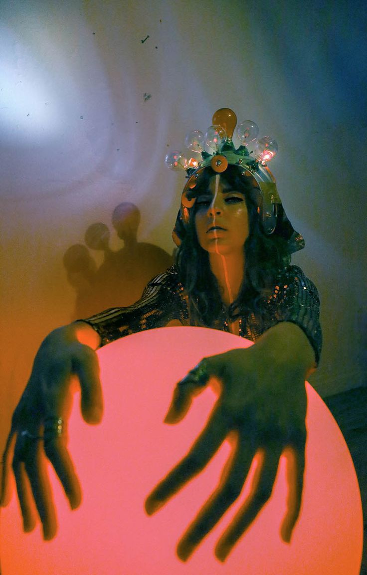 a woman with a futuristic headpiece of lightbulbs looks directly at the camera. she is holding a large, glowing orb.  the image is dark and has a surreal quality to it.  the woman's expression is mysterious and the lighting casts interesting shadows.