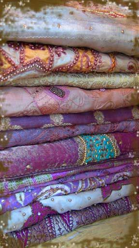 a stack of intricately woven fabric, likely saris, adorned with beautiful embroidery and beads. the fabrics are layered with varying colors, primarily purple, pink, white, and blue, creating a stunning visual spectacle. the delicate patterns and ornate embellishments on the fabric showcase the rich cultural heritage and artistry of indian and asian textile traditions. the image captures the beauty and elegance of traditional clothing and the intricate craftsmanship involved in their creation.