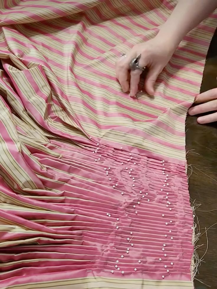 a closeup shot shows hands pinning pleats into striped pink and beige fabric. the fabric is neatly folded into narrow pleats and secured with numerous sewing pins, while the upper portion of the fabric lays flat. the person's hands are visible, with a large ring on one hand.